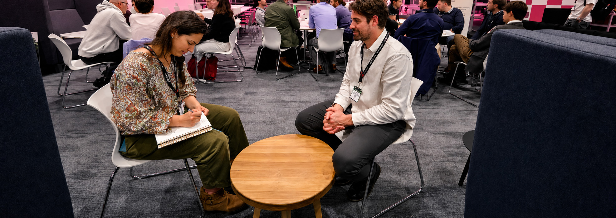 Two people sit across from each other at a small wooden table during a Futurebuild networking session, with a pink-branded conference background.
